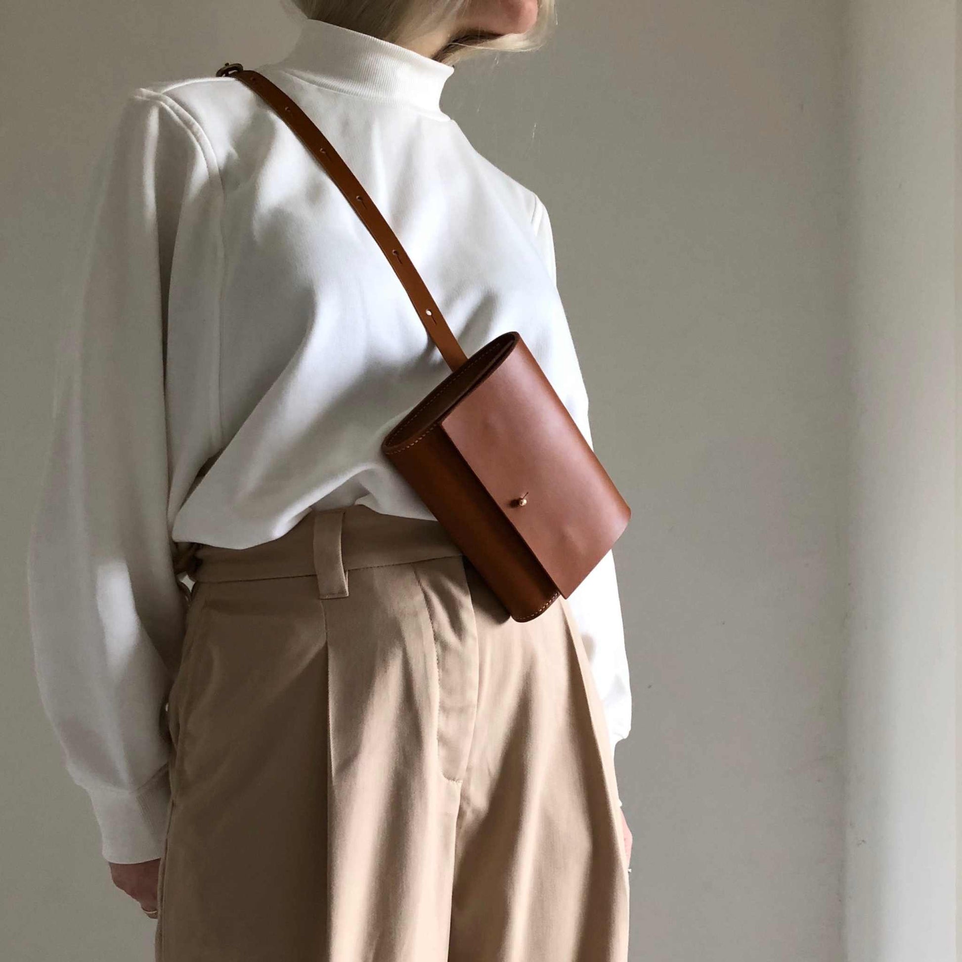 Tan leather belt bag worn crossbody over a white shirt and beige pants.