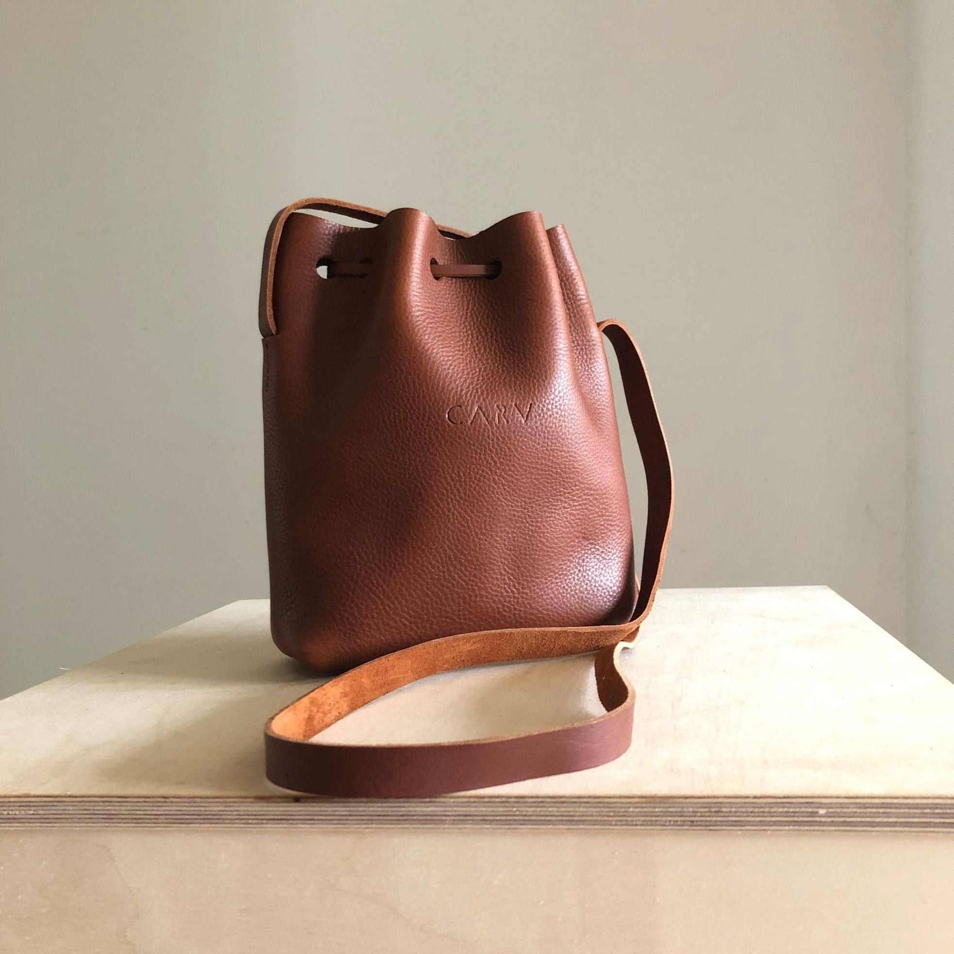 Nomad Mini Leather Bucket Bag in tan brown with adjustable strap, handcrafted from vegetable-tanned leather, featuring a raised grain texture.