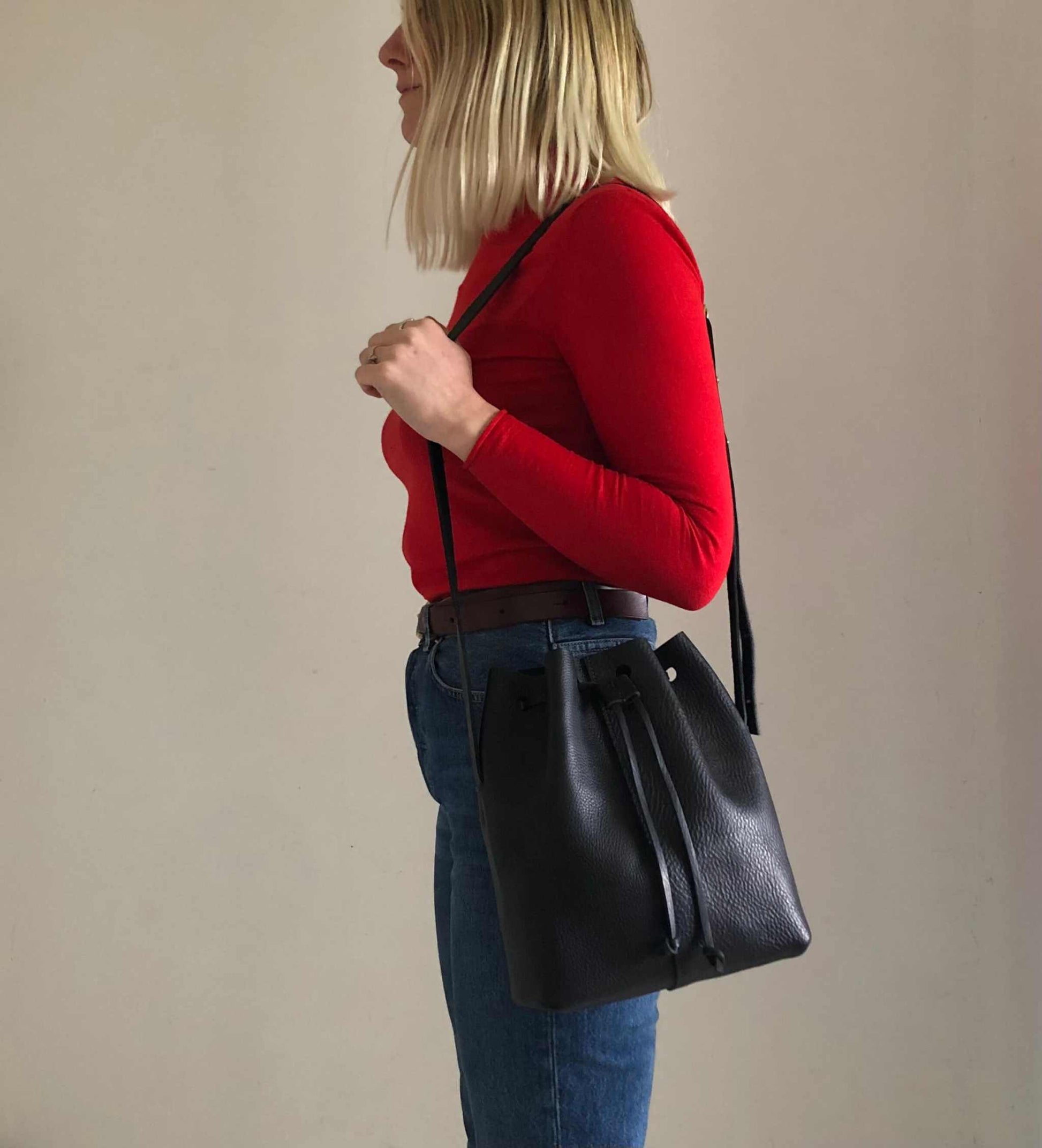 Nomad Midi Tan Leather Bucket Bag with adjustable strap, worn crossbody by a person in a red top and jeans.