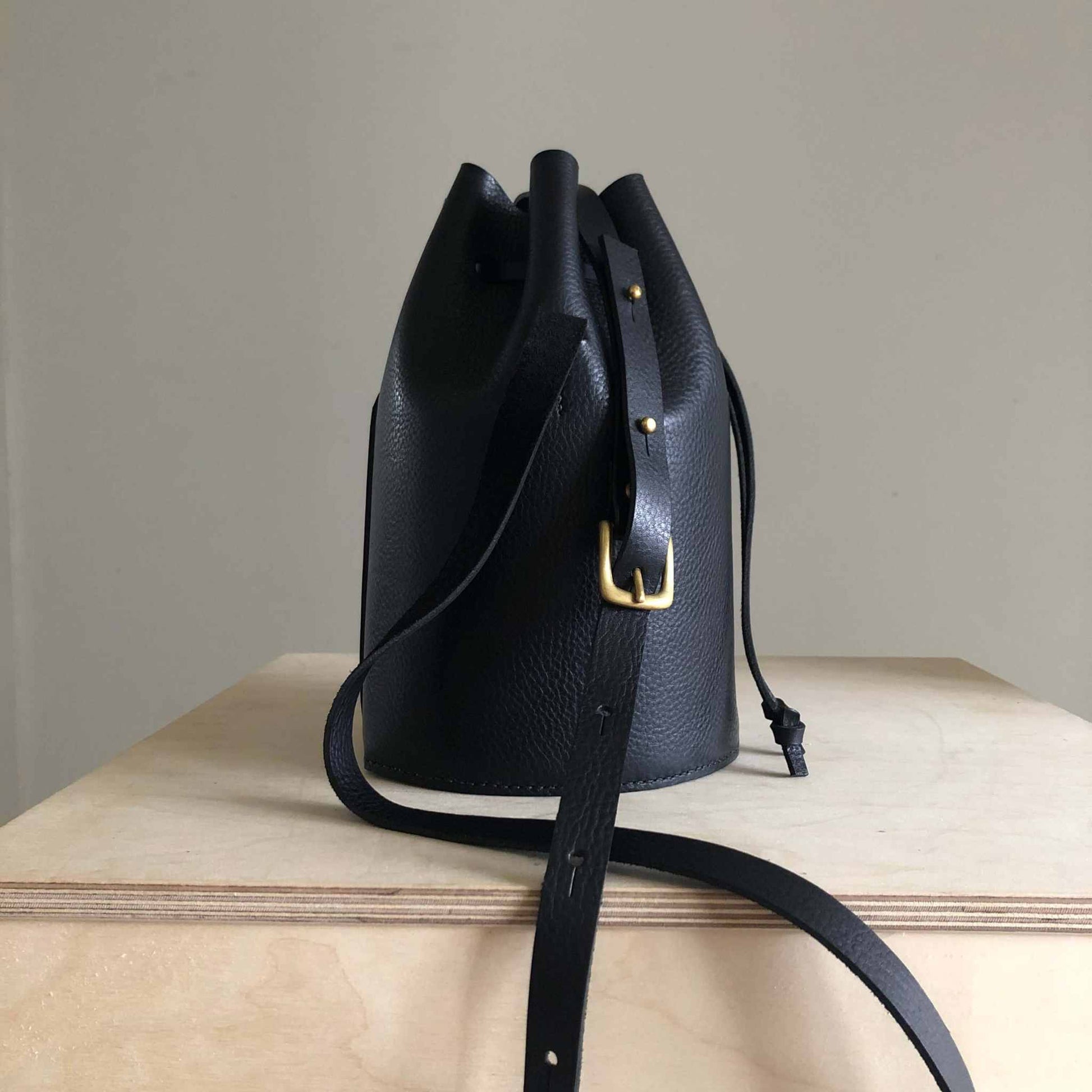 Black cylinder leather bucket bag with adjustable strap and brass buckle on wooden surface.