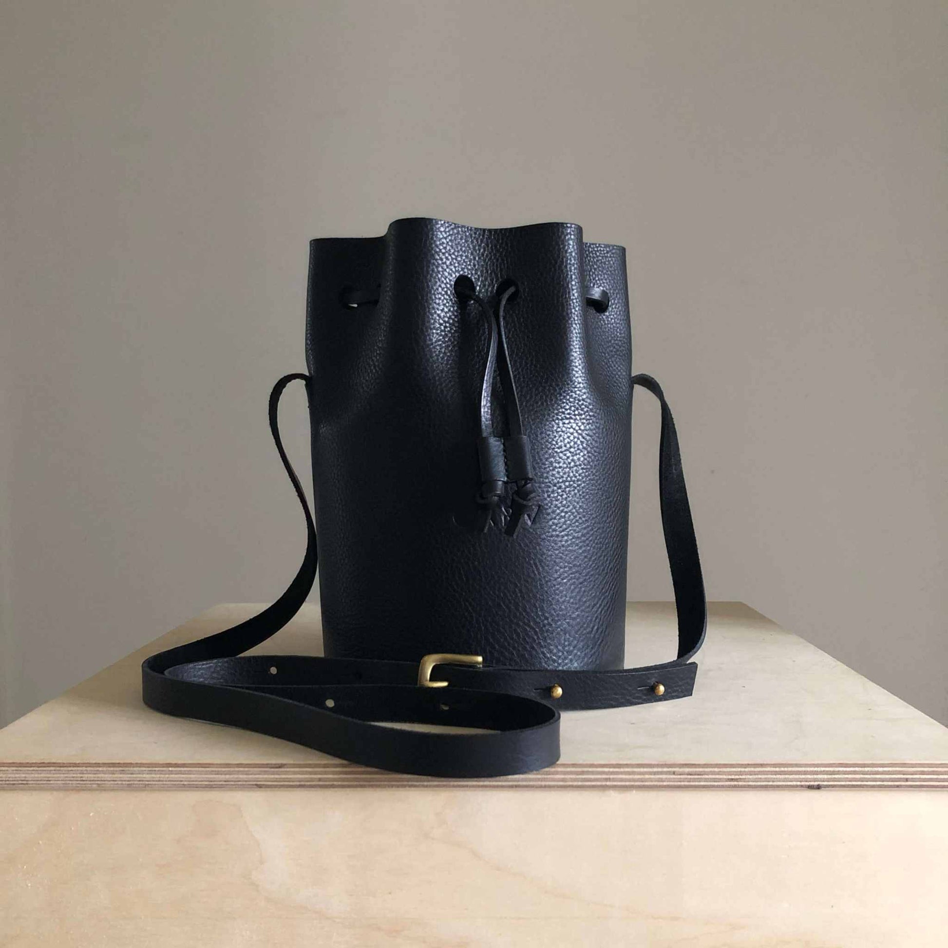 Cylinder Leather Bucket Bag in black with adjustable strap and solid brass buckle on display.