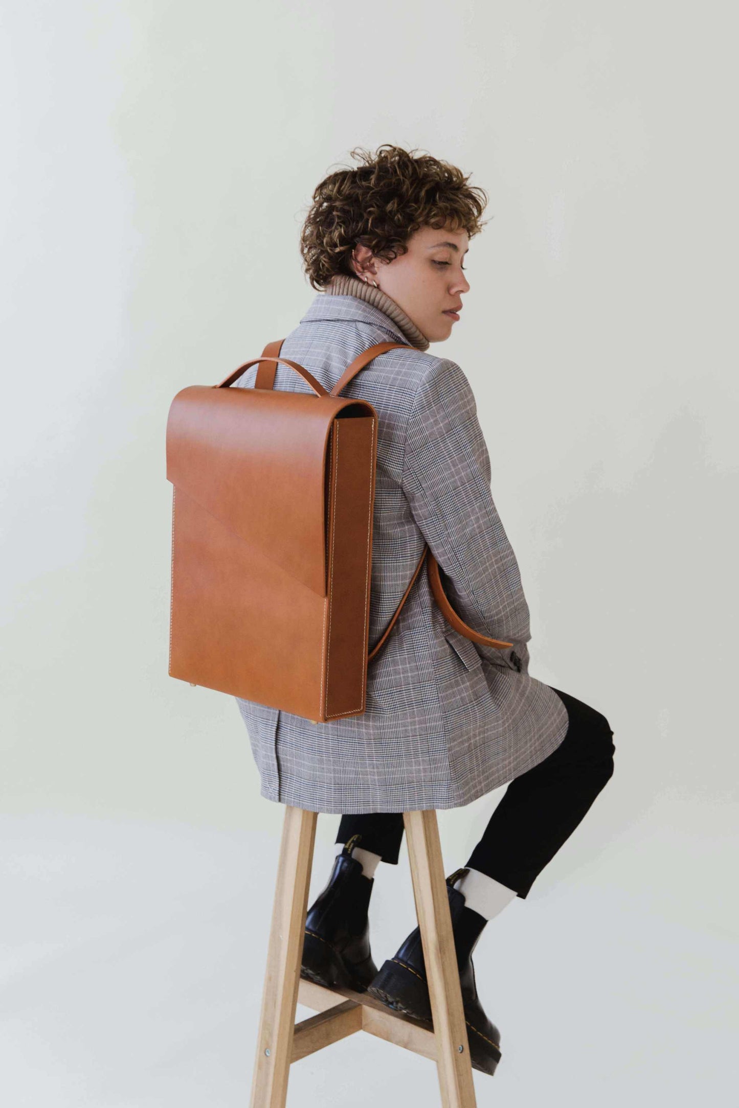 Minimalist tan leather backpack on a person wearing a gray plaid coat, seated on a stool.