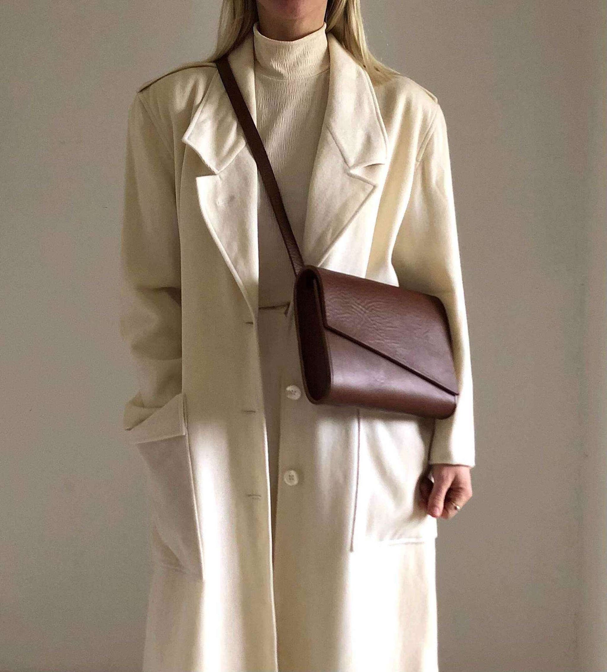 Tan leather crossbody bag with asymmetrical flap worn by person in cream coat.