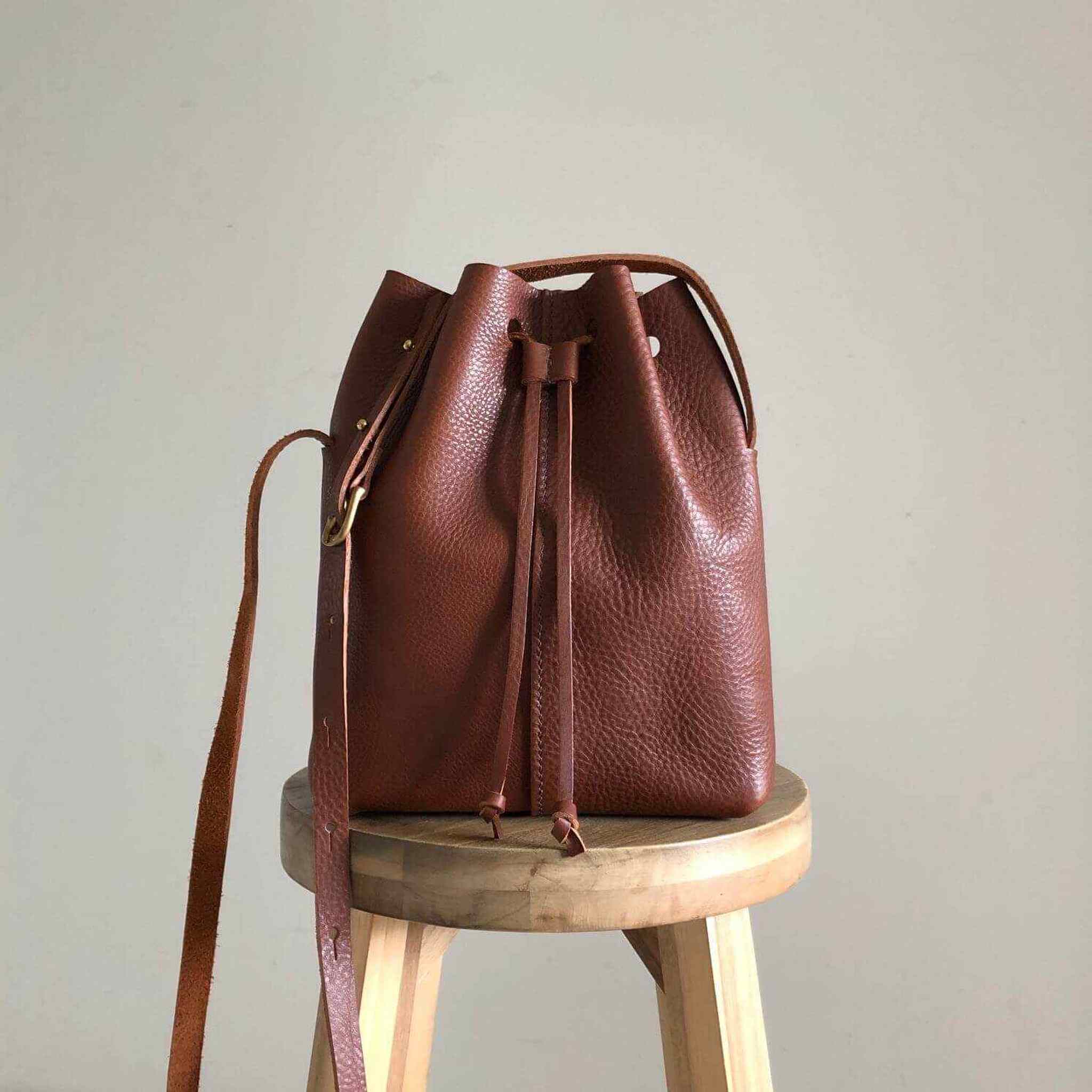 Nomad Midi Tan Leather Bucket Bag on stool, adjustable strap, minimalist design.