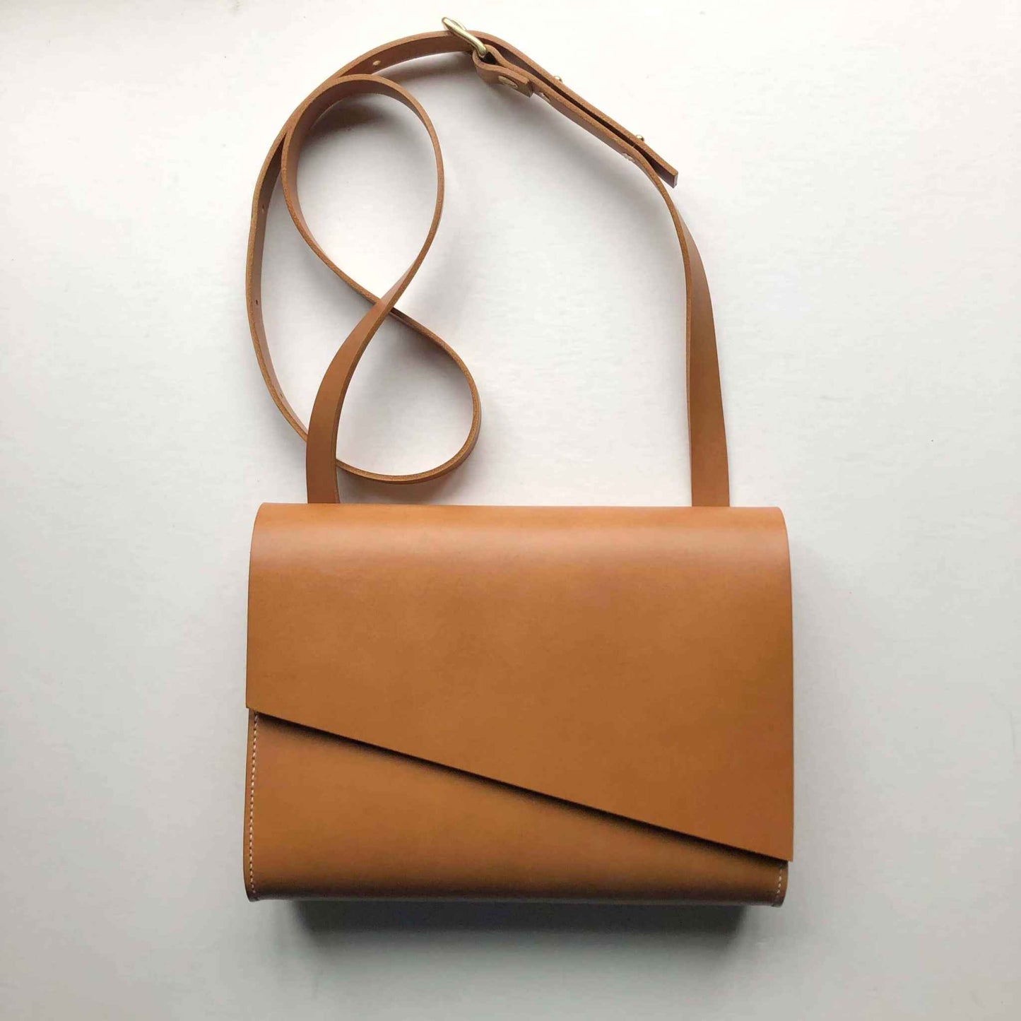 Tan leather crossbody bag with adjustable strap and asymmetrical flap design.