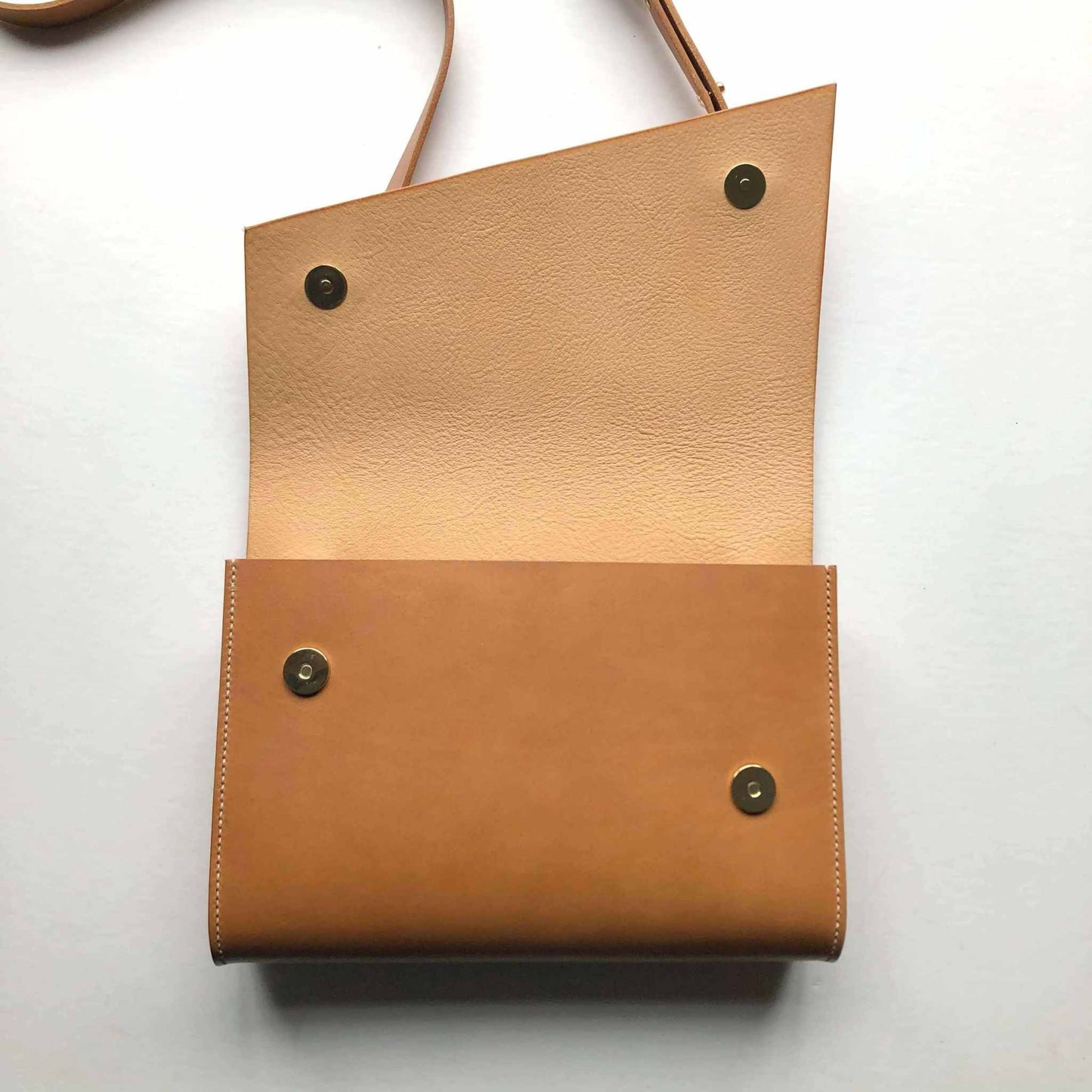 Tan leather crossbody bag with an asymmetrical flap and brass snap closures, featuring adjustable strap.