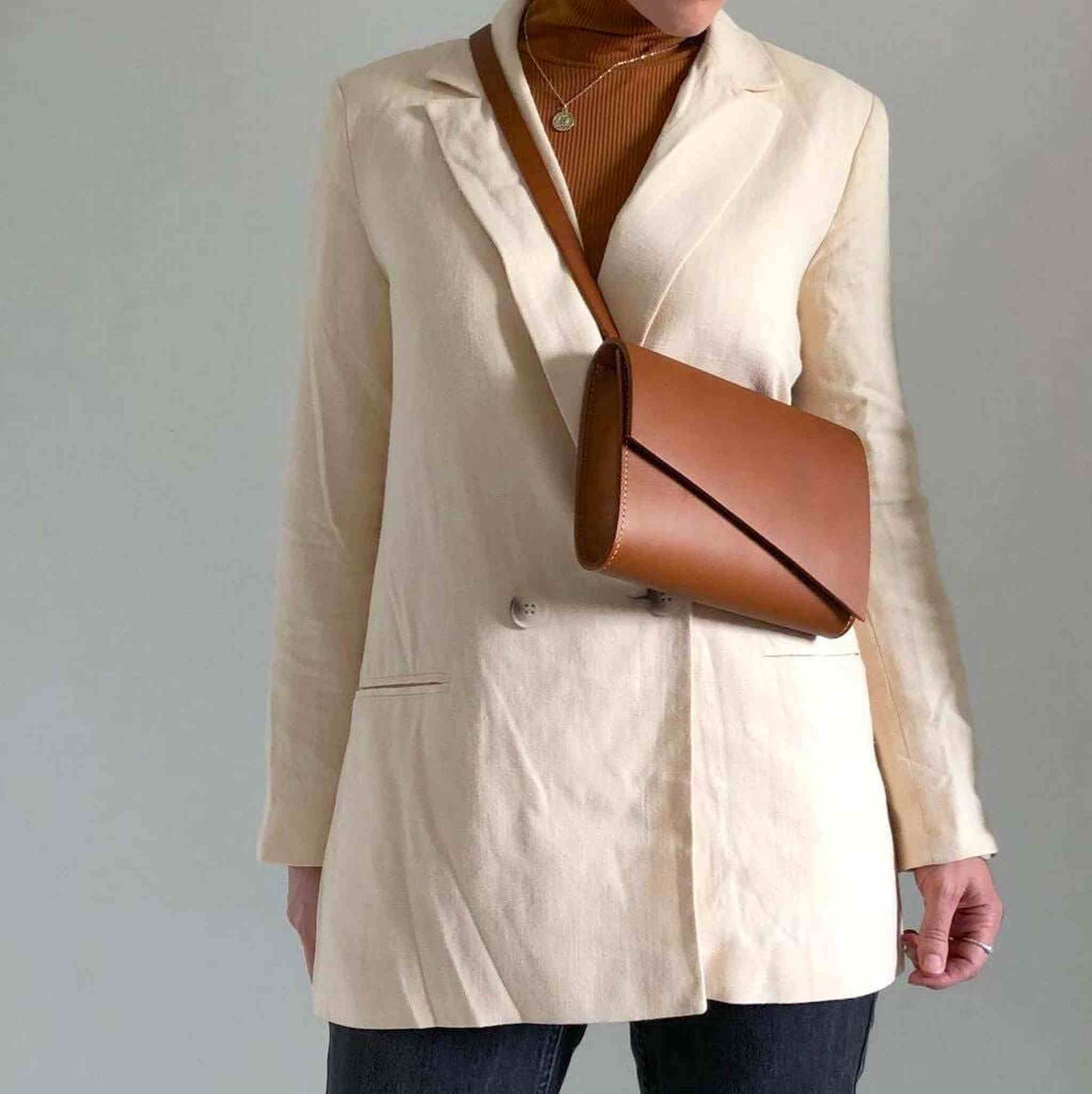 Tan leather crossbody bag worn over cream blazer, showcasing minimalist design and adjustable strap.