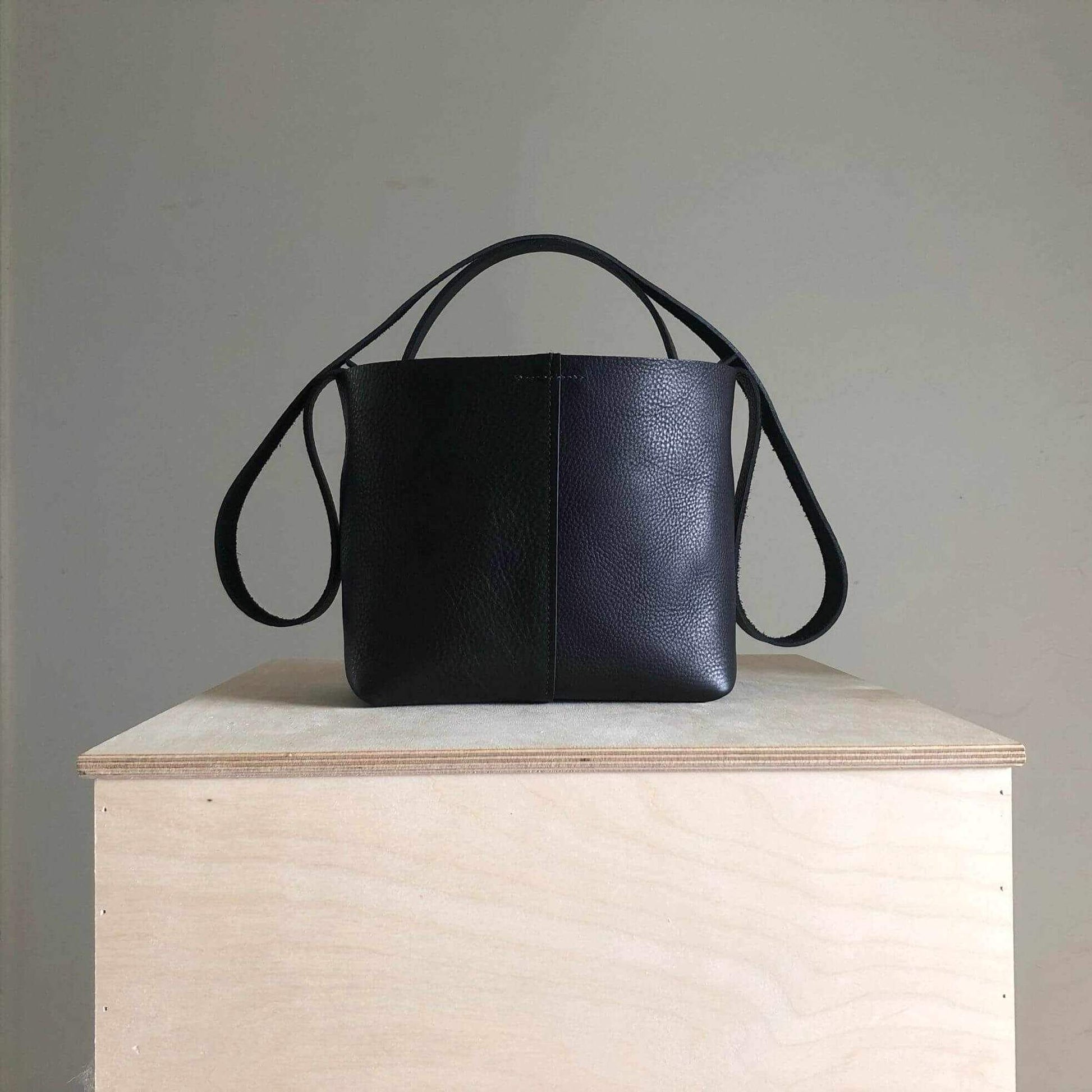 Basket Mini Leather Tote Bag by CARV in black displayed on wooden surface, showcasing its modern basket silhouette and interchangeable straps.