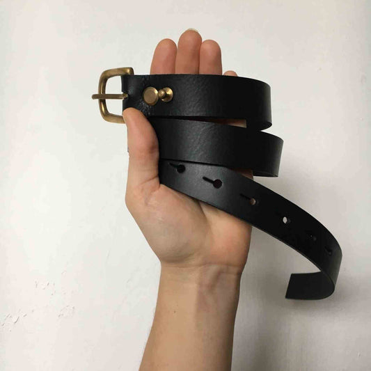 Nomad Midi Width Black Leather Belt with brass buckle held in a hand.